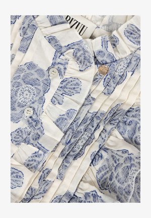 Biały materiał z niebieskim fakturowanym kwiatowym nadrukiem, pionowymi plisami, białymi guzikami, falbankami na krawędziach oraz małą okrągłą metalową metką z nazwą marki.