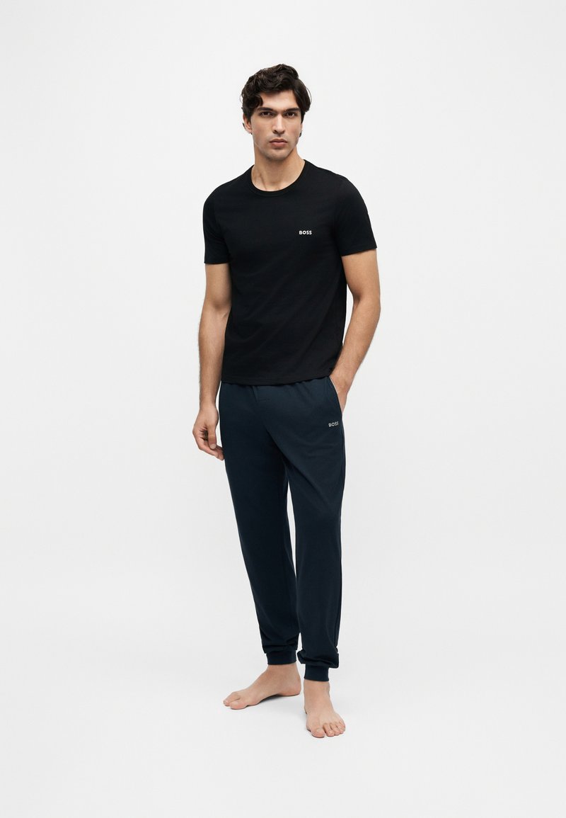 Mann steht barfuß, trägt ein schwarzes T-Shirt mit kurzen Ärmeln und dunkle Jogginghosen mit "BOSS"-Logos auf beiden Seiten.