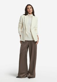 Elfenbeinfarbener, gerippter Blazer über einem hellen Sweater, kombiniert mit weiten, braunen Satin-Hosen. Der Blazer hat eine taillierte Passform und einen Reverskragen.