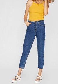 Modré jeans s vysokým pasem a rovnými nohavicemi, s jemnými švy, nošené s žlutým žebrovaným topem a bílými sandály.