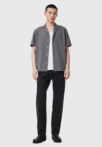 Camisa gris de manga corta con cuello tipo notch, cierre delantero de botones, combinada con jeans negros. Modelo lleva una camiseta blanca debajo. Calzado negro.