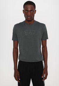 Temno siva bombažna majica s kratkimi rokavi, ki na sprednji strani vsebuje ton-na-ton logotip EA7 Emporio Armani. Okrogel izrez, preprost dizajn.