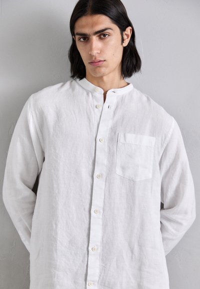 Camicia bianca in lino con colletto alla coreana, chiusura con bottoni sul davanti e taschino sul lato sinistro del petto. Il tessuto ha una finitura texturizzata.