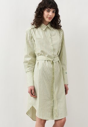 Kobieta o kręconych brązowych włosach, ubrana w jasnożółtą sukienkę koszulową z pionowymi zielonymi paskami oraz pasującym paskiem, stojąca na jednolitym tle.