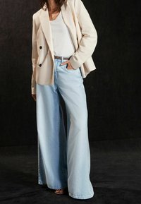 Blazer beige clair à manches longues, associé à un jean en denim bleu clair taille haute et à jambes larges. Débardeur blanc uni en dessous.