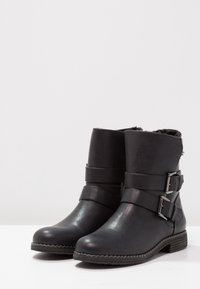 Botas negras hasta el tobillo hechas de cuero sintético con forro de piel sintética. Presentan dos correas ajustables con hebillas plateadas y suelas texturizadas.