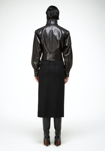 Žena stojaca chrbtom, oblečená v čiernej koženej bunde s vysokým golierom, čiernej midi sukni s vreckami na zadnej strane a čiernych čižmách na vysokom podpätku.