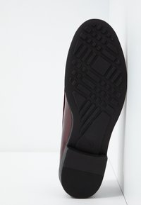 Semelle d'une chaussure habillée noire inclinée contre un mur blanc d'angle, montrant une semelle extérieure en caoutchouc noir texturée avec des motifs géométriques.