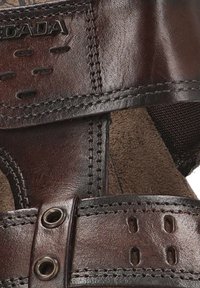 Sandalia de cuero marrón con tiras cosidas, acentos de hardware metálico y un forro similar a gamuza. Incluye detalles perforados en las tiras.