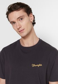Camiseta de algodón negra con cuello redondo, que presenta un pequeño logo bordado en amarillo de "Wrangler" en el lado izquierdo del pecho. Mangas cortas.