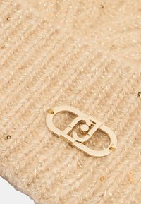 Cappello beige lavorato a maglia con un motivo testurizzato, dettagli metallici dorati e un logo ovale centrale. Materiale morbido con una leggera lucentezza.