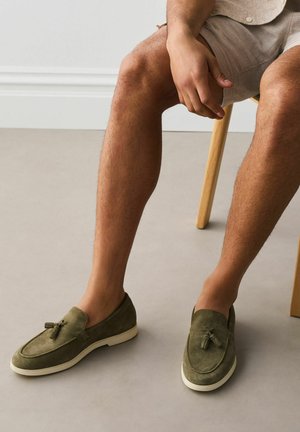 Homme assis sur une chaise en bois, portant des mocassins en daim vert à glands et un short beige, devant un fond uni.