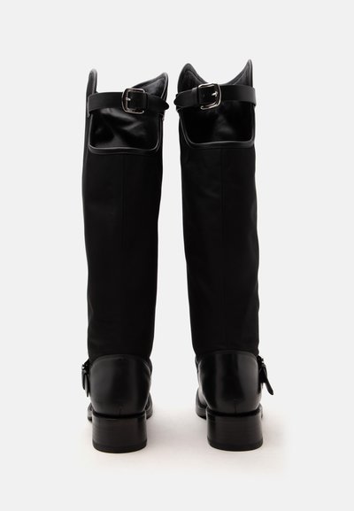 Paire de bottes hautes en cuir noir avec des sangles à boucle en haut et autour de la cheville, vues de dos sur fond blanc.