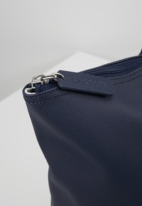 Sac à main en nylon texturé bleu marine avec une fermeture éclair et une étiquette Lacoste. Forme arrondie avec des détails de couture fines.