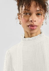 Weiße Bluse mit hohem, gerafftem Kragen, vertikalen Nadelstreifen und strukturierten Punkten. Kurze Ärmel mit einem zarten, weichen Finish.