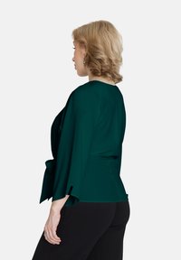 Zelená blúzka s elegantnými, rozšírenými rukávmi a peplum pásom. Hladká textúra, s jednoduchým dizajnom a kontrastnými čiernymi akcentmi.