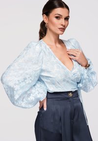 Femme aux cheveux foncés attachés en queue de cheval, portant un chemisier bleu clair à manches bouffantes fleuries et un pantalon en satin foncé taille haute, accessoirisée de boucles d'oreilles et d'un bracelet en or.