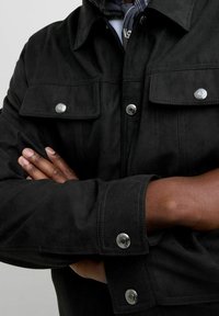 Czarna zamszowa kurtka z dwoma kieszeniami na piersi, srebrnymi zatrzaskami i zapinanym mankietem. Widoczny jest złożony kołnierz oraz fakturowana tkanina.