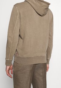 Felpa con cappuccio marrone realizzata in tessuto morbido, con polsini e orlo a coste, caratterizzata da un design semplificato e dettagli minimi.