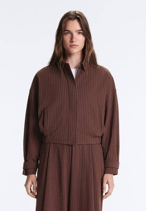 Femme portant une veste marron à fines rayures avec une fermeture éclair et un pantalon large assorti, debout sur un fond blanc.