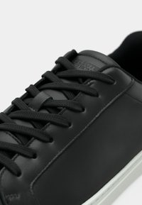 Baskets en cuir synthétique noir avec des lacets plats noirs, une texture lisse et un logo discret sur la tige. Semelle blanche visible en bas.