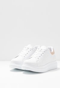 Bílé tenisky s hladkým koženým svrškem, kulatou špičkou a mírně vyvýšenou bílou podrážkou. Na patě mají světle hnědý akcent.