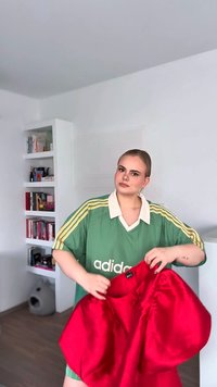 Zelenožltý pruhovaný nadrozmerný sveter s bielym golierom; drží jasne červený, mierne pokrčený kúsok oblečenia. Minimalistické biele pozadie.