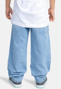 Světle modré džíny s širokými nohavicemi, se zadní kapsou a zahnuta hemem. Nošeny s šedými teniskami a bílým tričkem.