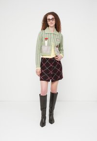 Νεαρή γυναίκα με σγουρά μαλλιά φοράει πράσινη ριγέ ζακέτα με λουλουδένια και γατοειδή μπαλώματα, μπορντό καρό φούστα και μαύρες μπότες μέχρι το γόνατο.