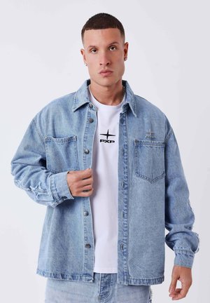 Homme aux cheveux courts portant une veste en jean bleu clair par-dessus un t-shirt blanc avec un logo noir, debout devant un fond clair uni.