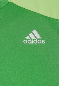 Zelená atletická látka s hladkou texturou a stříbrným logem Adidas na levé straně. Materiál působí lehce a prodyšně.