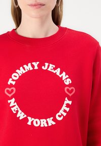 Rød hettegenser laget av bomull, med et rundt grafisk trykk som har "TOMMY JEANS" og "NEW YORK CITY" i fete hvite bokstaver, pyntet med hjerter.