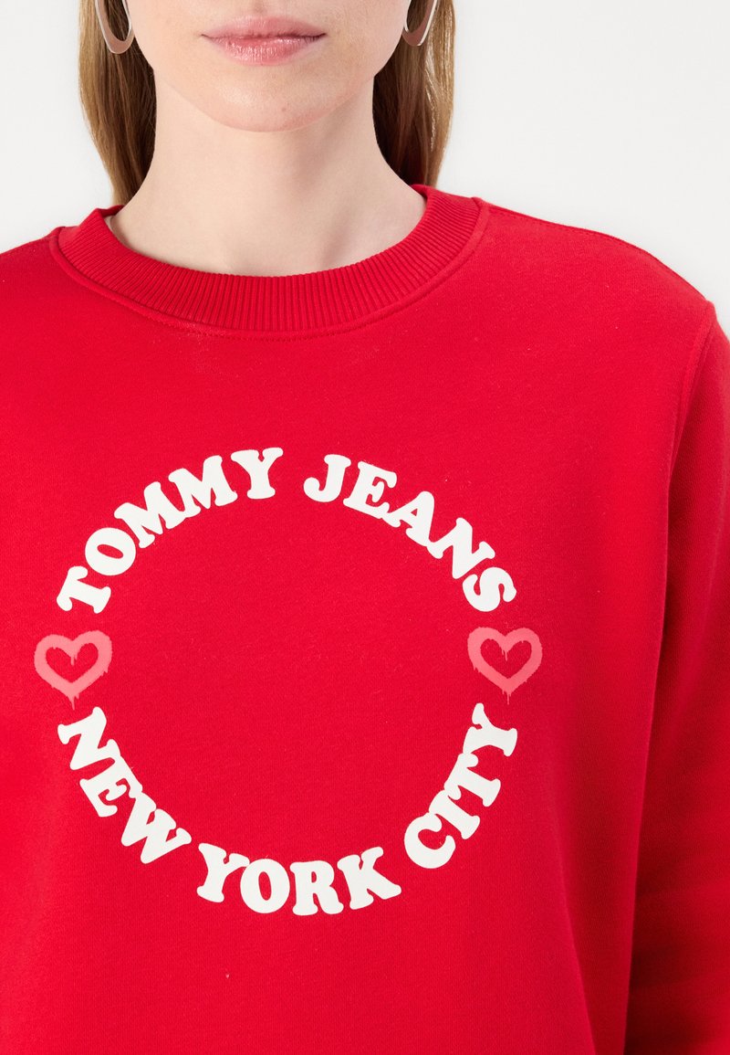 Felpa rossa in cotone, con una grafica rotonda che presenta "TOMMY JEANS" e "NEW YORK CITY" in bold lettere bianche, accentuata da cuori.