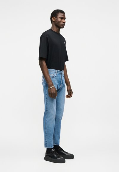 Homme debout de profil portant un t-shirt noir oversize, un jean bleu clair, des baskets noires et un bracelet de perles au poignet gauche.