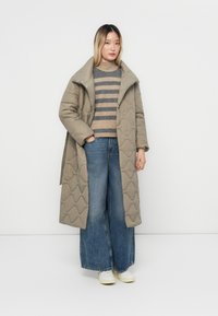 Vrouw in een gewatteerde lange beige jas, gestreepte bruin en grijze trui, wijde blauwe jeans en witte sneakers, staand tegen een witte achtergrond.
