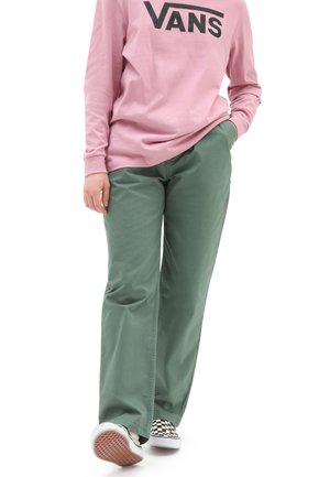 Persona che indossa un felpa rosa Vans, pantaloni verdi e scarpe slip-on a scacchi, mentre cammina con una mano in tasca.