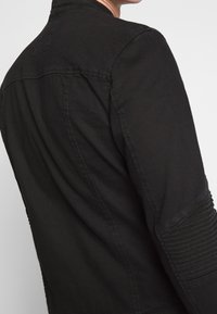 Veste noire en tissu mat, avec des nervures texturées sur les épaules, un col droit et un design minimaliste.