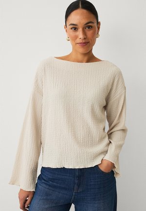 Frau mit dunklem Haar und goldenen Ohrringen, die eine strukturierte, cremeweiße Bluse mit langen Ärmeln und blaue Jeans trägt, steht mit einer Hand in der Tasche.