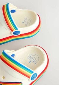 Baltas gumijas klogas ar varavīksnes krāsas siksniņu zilā, zaļā, dzeltenā, oranžā un sarkanā krāsā. Logotipā ir zilas krāsas apaļa detaļa. Redzama teksturēta zolīte.