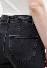 Άτομο που φοράει μαύρο τζιν από denim με μεγάλη πίσω τσέπη και μαύρη δερμάτινη ετικέτα στη ζώνη, σε συνδυασμό με λευκό τοπ.