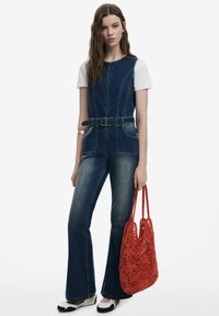 Denim jumpsuit with a belted waist, flared legs, dark blue color, front pockets, paired with a red crochet tote bag and black-and-white shoes.