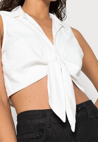 Camicetta bianca corta realizzata in tessuto morbido, con chiusura a fiocco frontale, colletto e design senza maniche. Indossata con pantaloni neri.