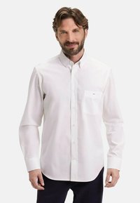 Homme aux cheveux bruns et à la barbe, vêtu d'une chemise blanche à manches longues et de pantalons foncés, se tenant devant un fond clair uni.