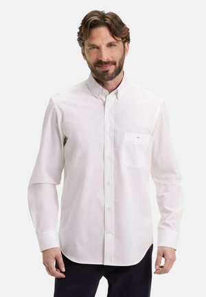 Homme aux cheveux bruns et à la barbe, vêtu d'une chemise blanche à manches longues et de pantalons foncés, se tenant devant un fond clair uni.