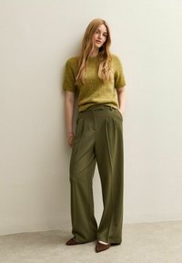 Suéter de punto de manga corta en verde oliva combinado con pantalones de lino de tiro alto y pernera ancha en color oliva y zapatos marrones de punta. Texturas suaves visibles.
