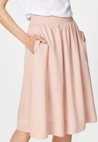 Nainen, jolla on ylhäältä vyötäröön nouseva, polvipituinen vaaleanpunainen hame, jossa on sivutaskut, ja valkoinen sisätaskuun laitettu paita.