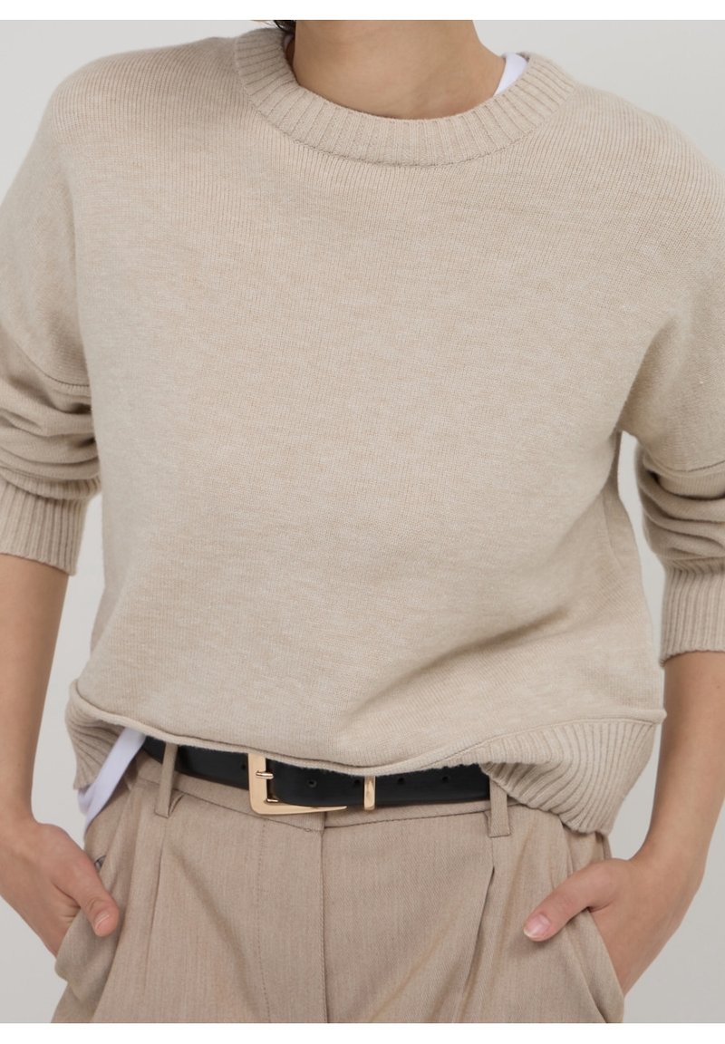 Personne portant un pull en maille beige rentré dans un pantalon beige avec une ceinture noire, mains dans les poches, et une chemise blanche qui dépasse à la taille.