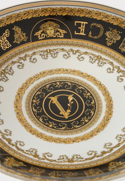 Διακοσμητικό λευκό πιάτο με πολύπλοκα μαύρα και χρυσά σχέδια, κεντρικό μονόγραμμα "V" και μοτίβο κεφαλής Μέδουσας στην άκρη.