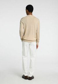 Μπεζ μακρυμάνικος πουλόβερ με ραβδωτά μανίκια, συνδυασμένο με λευκά παντελόνια και σκούρα παπούτσια. Το ύφασμα φαίνεται λείο με μια λεπτή υφή.