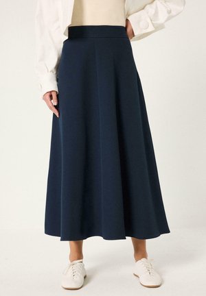 Γυναίκα φοράει μια μπλε navy μίντι φούστα σε σχήμα Α με ψηλή μέση, συνδυασμένη με λευκά αθλητικά παπούτσια και κρεμ πουκάμισο με μακριά μανίκια.